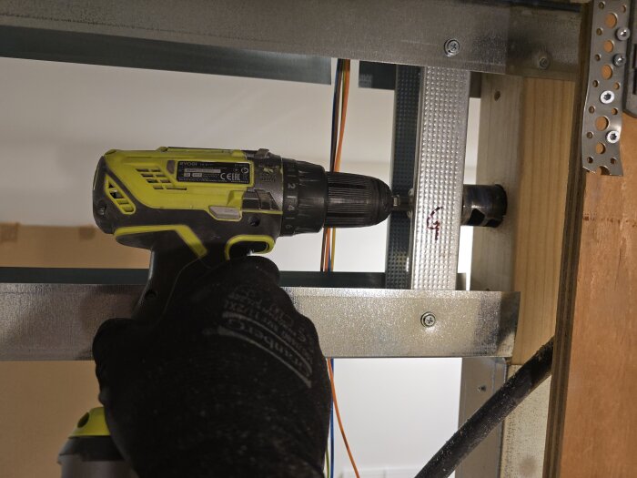 Hand med en Ryobi borrmaskin borrar i metallreglar för elinstallationer, synligt är färgade kablar och en träregel i bakgrunden.