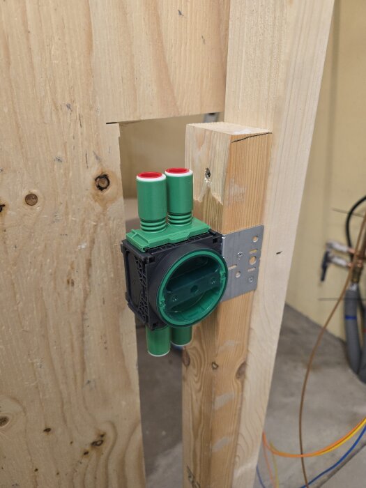 Väggmonterad eldosasockel i grönt fäst i träregel med metallbeslag och synliga ledningar i bakgrunden.
