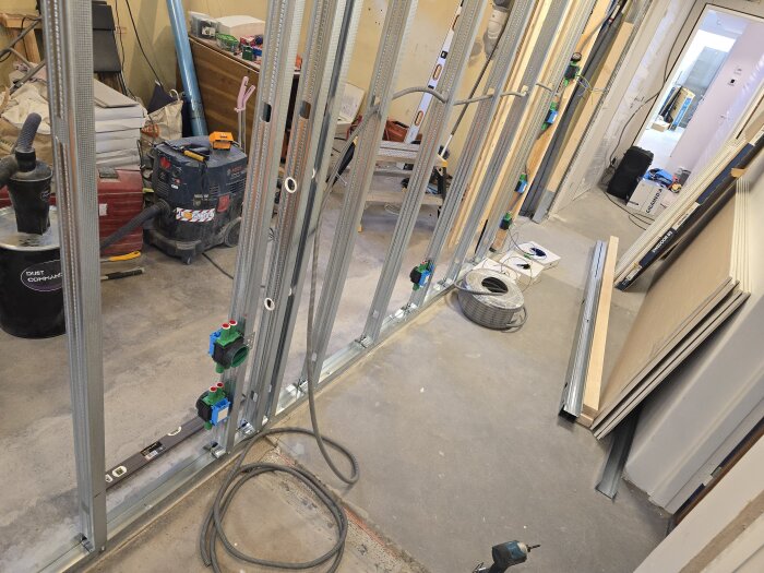 Byggarbetsplats med stålreglar och elinstallationer, pågående elektriskt arbete med sladdar och verktyg, och staplade byggmaterial i bakgrunden.