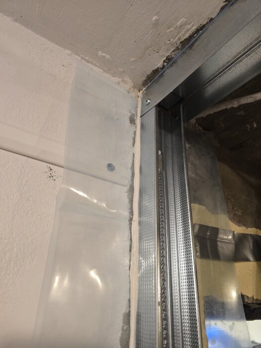 Närbild av ett hörn där två stålreglar möts i en vägg, med skruvar och plastfilm fastsatt mot väggen.