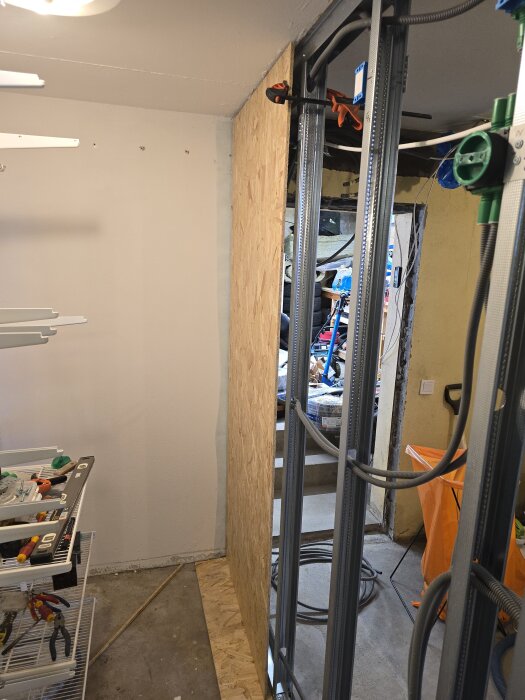 Byggarbetsplats med OSB-skiva och stålregelvägg under konstruktion. Flexslangar och elkablar syns på stålreglarna. Verktyg ligger på hyllor.