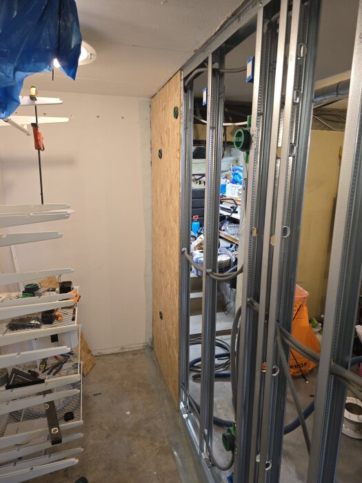 Byggarbetsplats med monterade stålskenor och OSB-skiva, hyllor och verktyg i förgrunden, rördragning synlig genom skenorna.