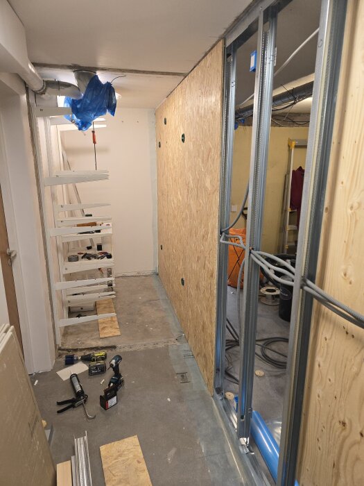 Korridor med monterad osb-skiva och synlig stålregelram för väggbygge, verktyg och material på golvet. Plaströr och hål för elinstallationer.