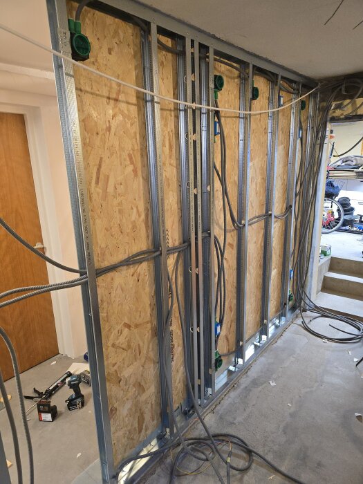 Väggkonstruktion med OSB-skivor monterade på stålreglar, synliga kablar dragna för elinstallation. Verktygssats ligger på golvet.