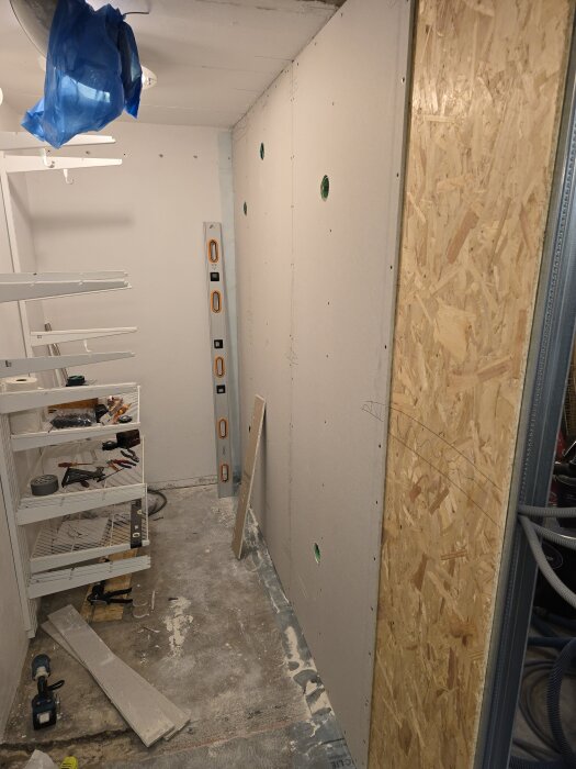 Renovering pågår med gipsskivor uppsatta på en vägg och verktyg och material spridda på ett betonggolv, hyllor till vänster.