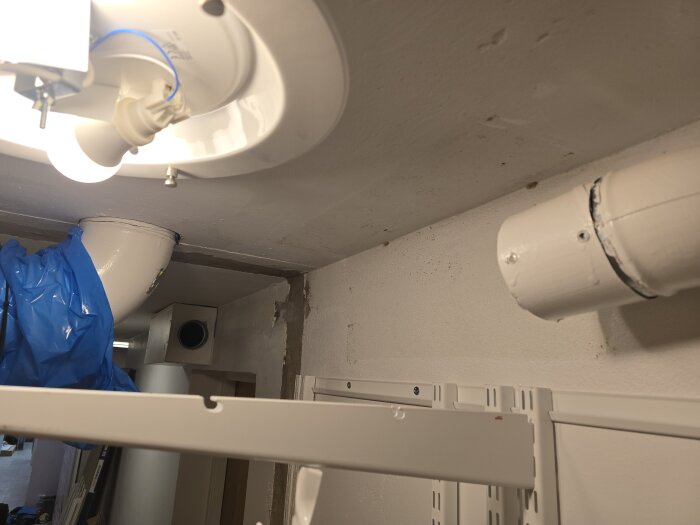 En belysningsarmatur i taket och ventilationsrör i ett rum under renovering med monterade skenor och blå plastpåse hängande.