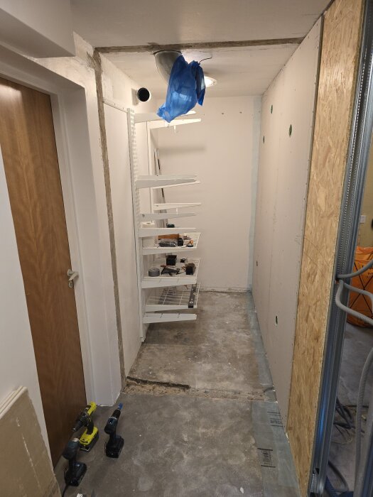 Renoveringsprojekt visar en smal hall med gipsväggar under konstruktion, hyllor längs väggen och verktyg på golvet, redo för byggarbete.