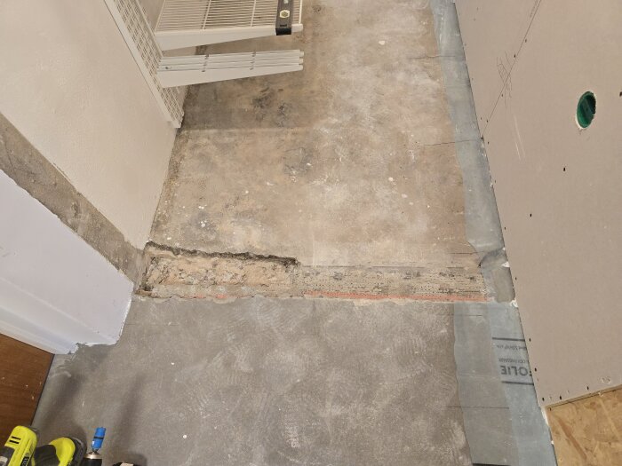 Byggprojekt: Rå golvyta med vägg av gipsmontering in progress, samt tydlig ledning för fuktbarriär med byggplast. Hyllplan och verktyg synliga.