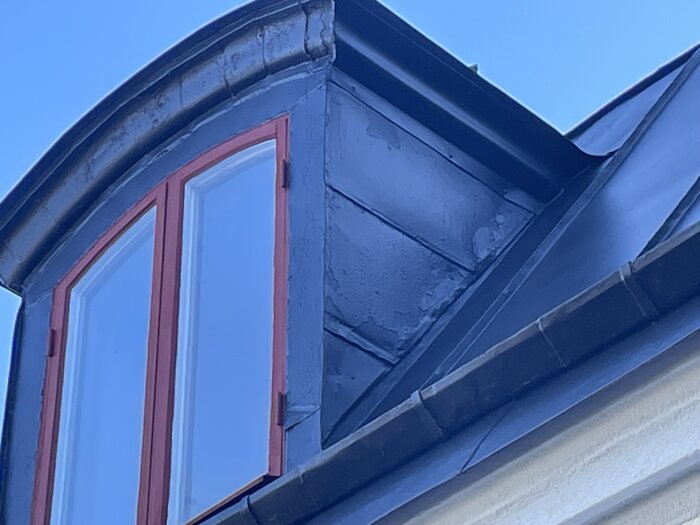 Ommålat plåttak med röd fönsterkarm och synlig färgavflagning, fotograferat under klar himmel.
