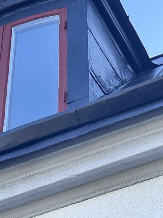 Ommålat plåttak med grå färg och röd fönsterkarm, detalj av husfasad och tak på nära håll.