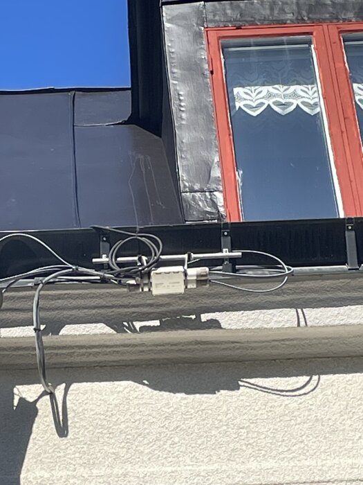 Plåttak med målat ytskikt och rött fönster, synliga kablar monterade på husfasaden under taket.