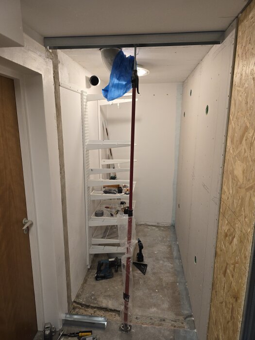 Renoveringsprojekt med gipsskivor, verktyg och hyllor i ett rum. En röd stolpe stöttar en takskena, och en blå plastpåse hänger i taket.