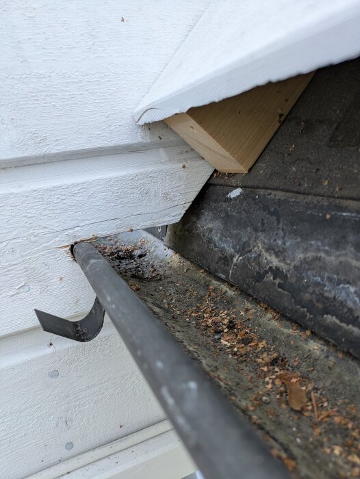 Närbild av en hängränna med anslutande reglar och takkonstruktion, fylld med löv och smuts. Plåtar och trä synliga vid anslutningen mot väggen.
