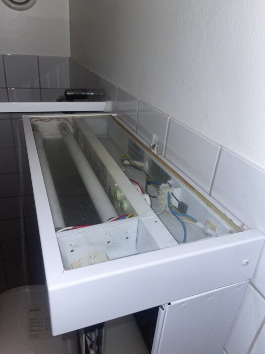Öppet badrumsskåp med synliga lysrör och elkablar, ingen synlig mekanism för att ta bort plexiglaset; vita kakelplattor i bakgrunden.