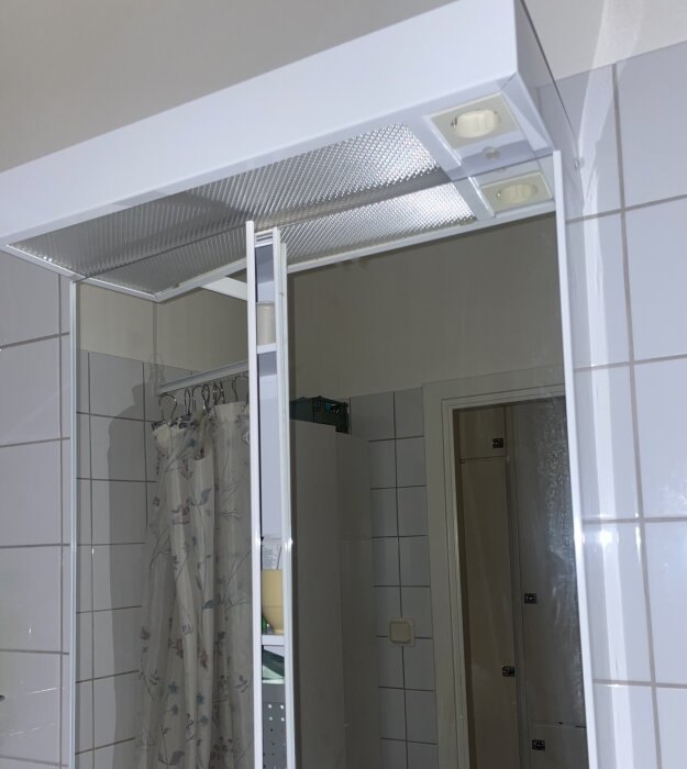 Badrumsskåp med plexiglas utan synliga skruvar, svårt att öppna för att byta lysrör. Vitt kakel i bakgrunden, del av duschdraperi syns.