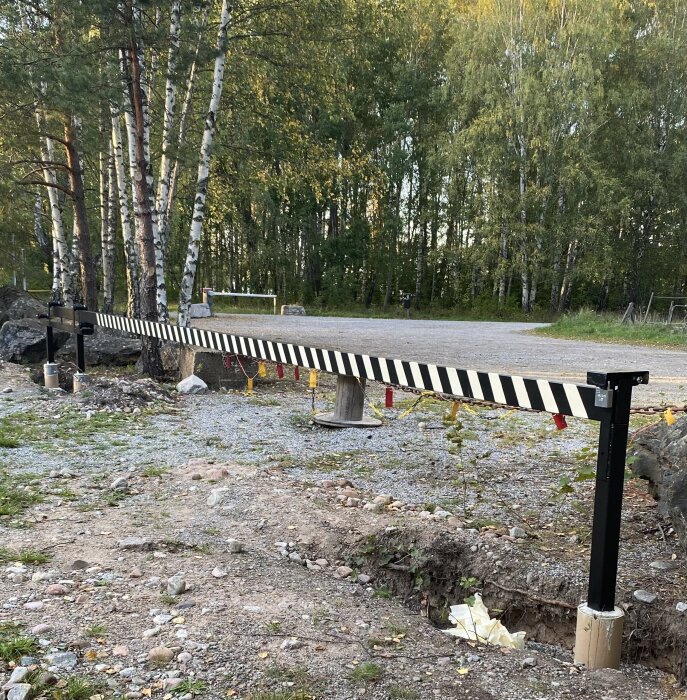 Svart- och vitrandig bom i naturmiljö, del av byggprojekt, står på grusig mark nära träd.