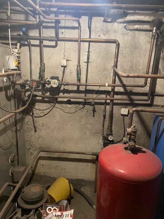 Rörsystem i källare med ett rött expansionskärl, rördragningar och en tryckmätare mot en grå betongvägg.