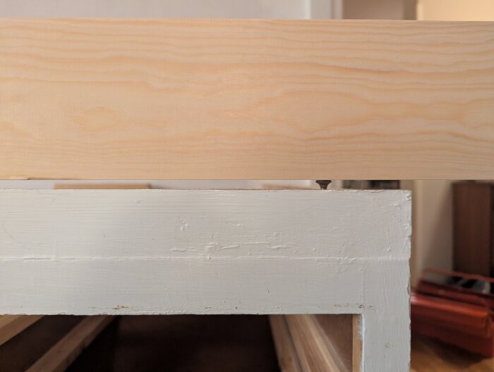 Närbild av träskiva ovanpå vitmålad skåpsram, med en synlig skruv mellan dem. Skåpskivan är något ojämn.