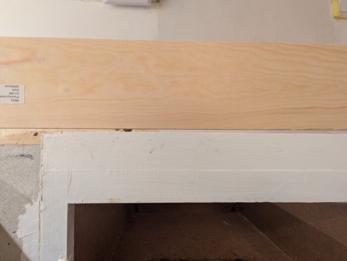 Träplanka placerad på en vitmålad sockel i ett kök, där målet är att justera nivåskillnad inför installation av diskmaskin.