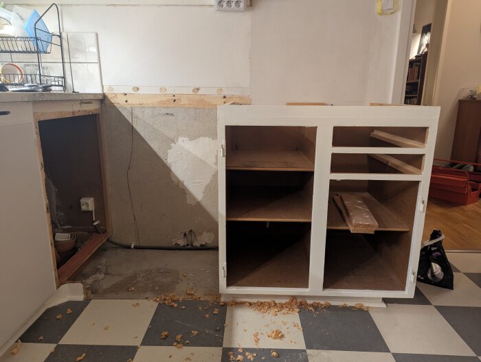 Tomt köksskåp på en svartvit rutig golv, omgiven av hyvelspån, med en öppning bredvid diskbänken för en planerad diskmaskin.