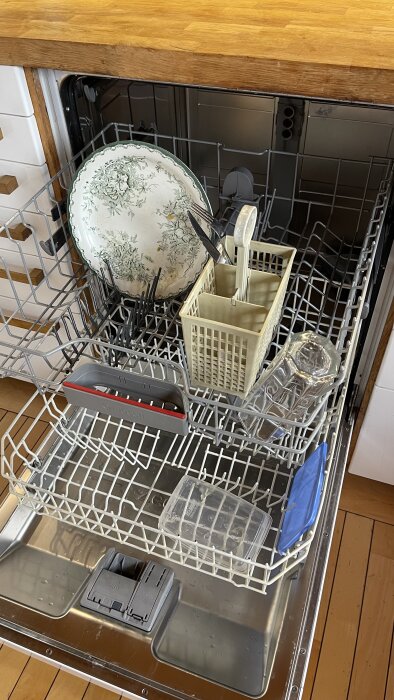 Öppen diskmaskin fylld med porslin och bestick, översta korgen med tallrikar och besticklåda, undre korg med plastruta och tomt utrymme.