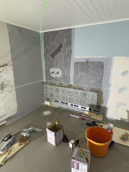 Kaklingsprojekt pågår med påbörjad kakelvägg. Laserlinje syns för att säkerställa perfekt avvägning. Verktyg och material utspridda på golv.