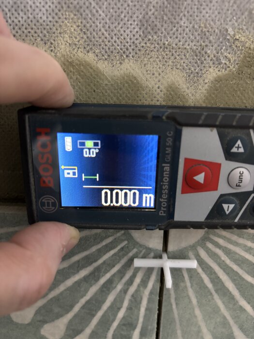 Digitalt vattenpass visar 0.0 grader vågrätt under kakling av vägg, med kakelkryss placerat mellan kakelplattor.