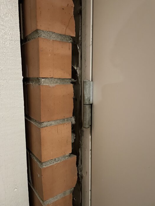 Spricka mellan tegelyttervägg och dörrkarm med exponerade fästen, där träreglar tidigare monterats över glipan.
