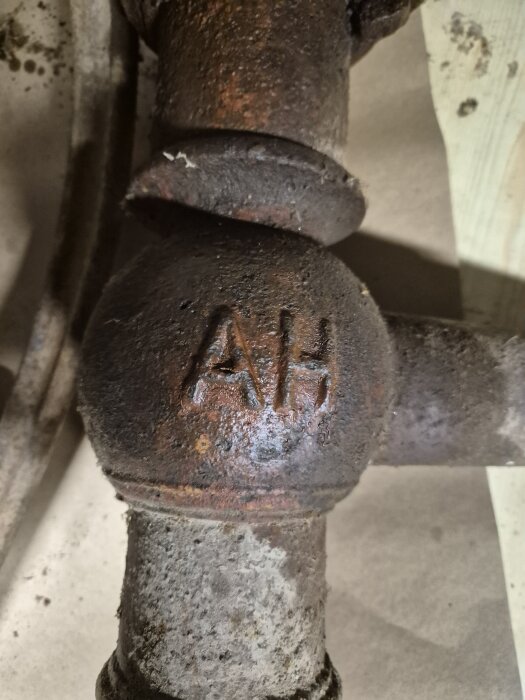Gårdspump i metall med bokstäverna 'AH' ingraverade, omgiven av rost och smuts.