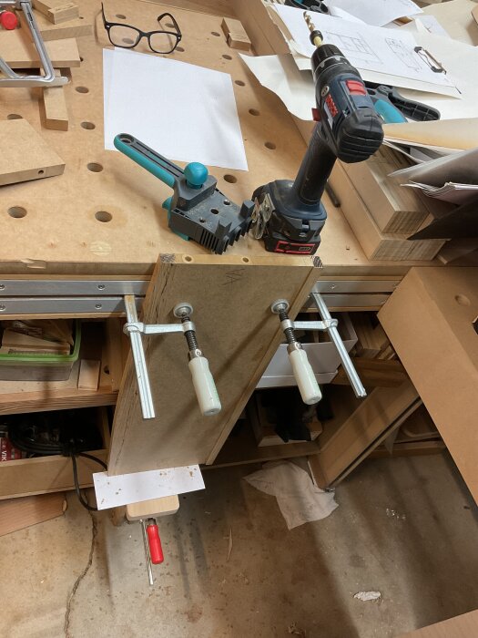 En arbetsbänk med träskiva fastspänd med tvingar, borrmaskin och jigg för plugghålsborrning, omgiven av verktyg och papper i verkstadsmiljö.