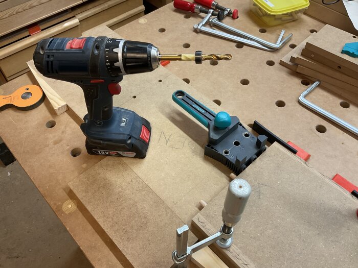 Borrmaskin och träpluggjigg på arbetsbänk med fastspända träbitar i verkstad. Verktyg och klämmor syns också.