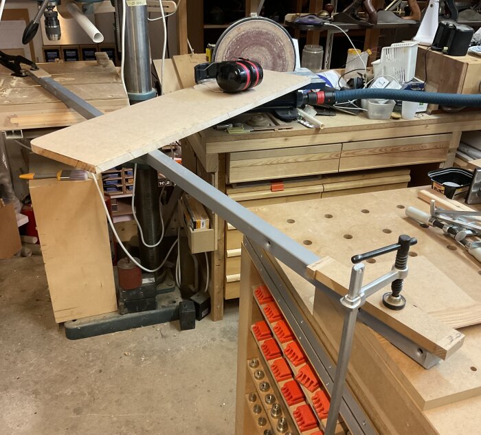 Verkstad med arbetsbänk, MDF-skiva med rundade hörn, hörselkåpor och slipmaskin; projekt pågår.