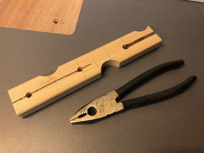 En hemmagjord träklämma ligger bredvid en kombinationstång på ett bord. Klämman har en slits för träpluggar.