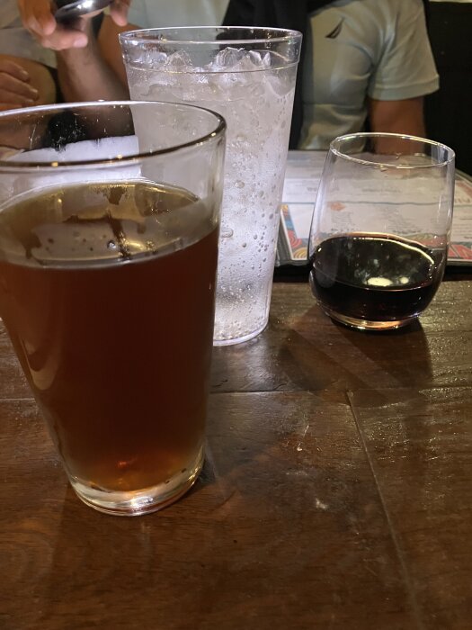 Glas med bärnstensfärgad öl, ett glas vatten med is och ett glas rött vin på ett träbord.