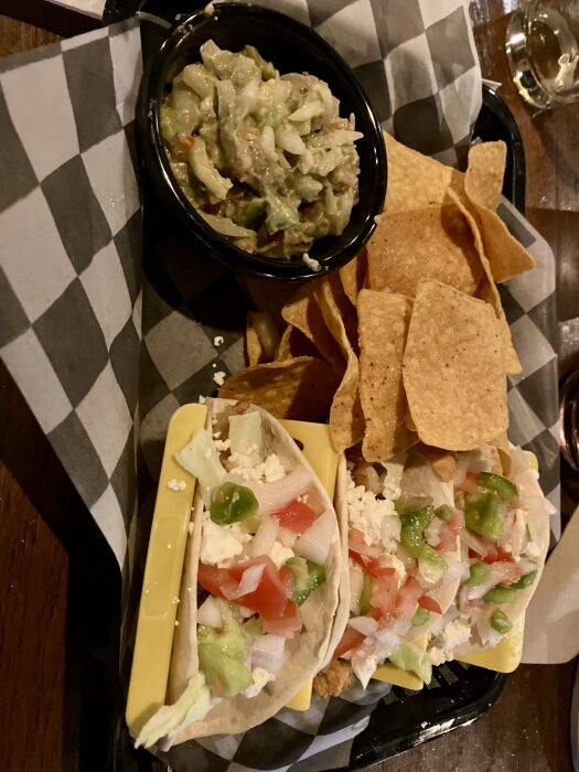 Shrimp tacos with diced vegetables, crumbled cheese, tortilla chips, and guacamole on a checkered paper tray.