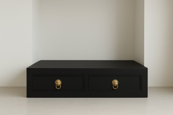 Svart sängbas med två lådor dekorerade med guldfärgade lejonhuvudhandtag, placerad mot en vit vägg.