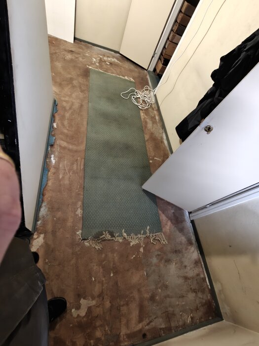 Ett källargolv med borttagen linoleummatta, exponerat betonggolv och en kvarvarande grön matta med fransar och hoprullad vit sladd.