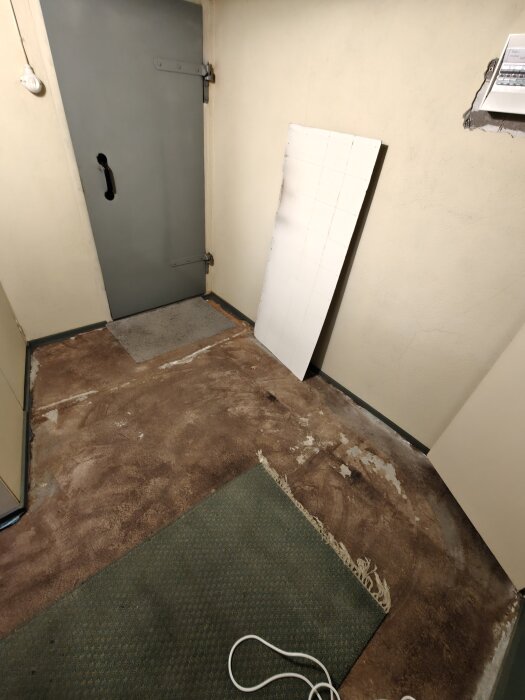 Källargolv med rester av gammal linoleummatta och betong synliga, grön trasmatta över en del av ytan, ståldörr i bakgrunden.