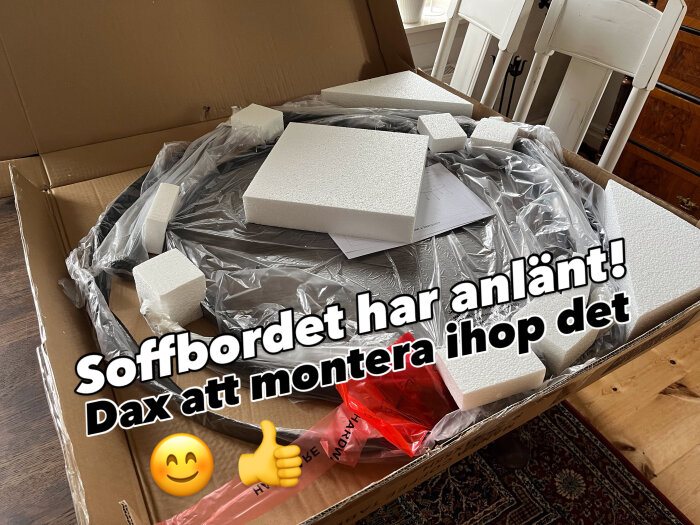 Runt soffbord i kartong redo att monteras. Omgivet av skyddande frigolit och plast. Text: "Soffbordet har anlänt! Dax att montera ihop det" plus emojis.