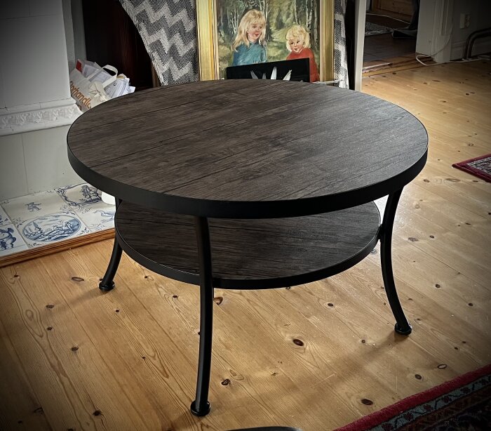 Runt soffbord i mörkt träliknande material med en hylla under, stående på ett trägolv med en tavla och tapetprover i bakgrunden.