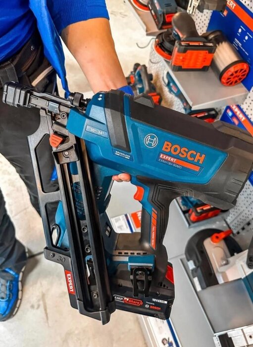 Spikpistol av märket Bosch Expert hålls i handen, omgiven av verktyg på en hylla i en butik.