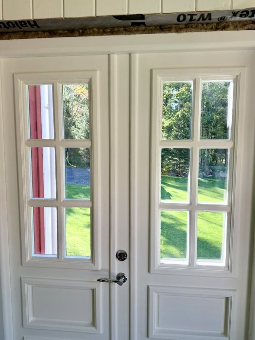 Vita dubbeldörrar med glaspartier, omgivna av grönska. Diskussion om kondensproblem mellan glaspartierna invändigt.