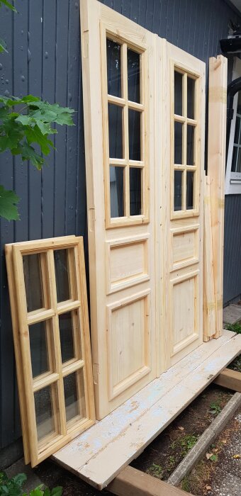 Nya träfönster- och dörrkarmar lutade mot ett gråmålat trävägg. En fönsterkarm står lutad bredvid dubbeldörrarna.