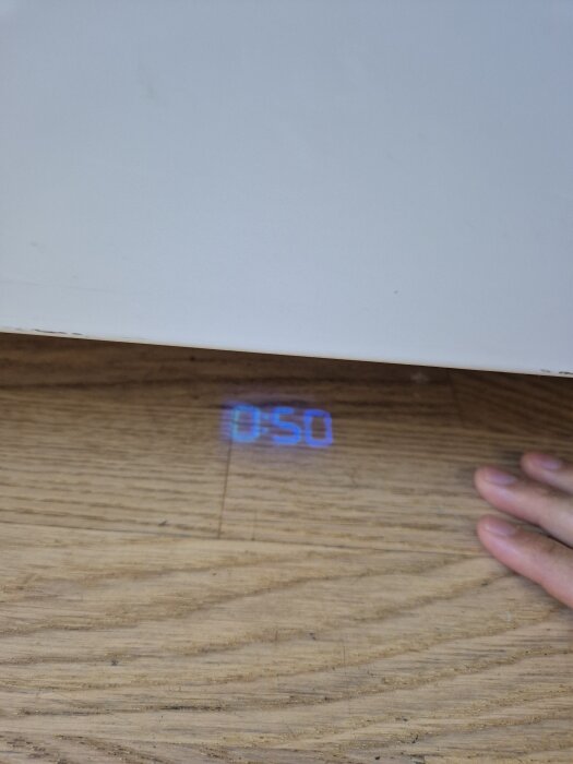 Projektion av siffror på golvet från en dold enhet under en hylla, med en hand bredvid.