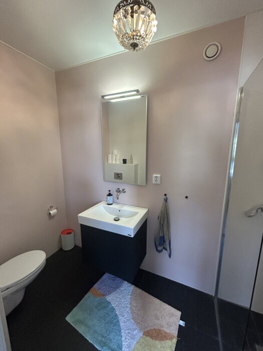 Gästtoalett med väggmonterad kran, spegel, handfat, dusch och pastellfärgade väggar. Det finns en taklampa och en färgglad matta.