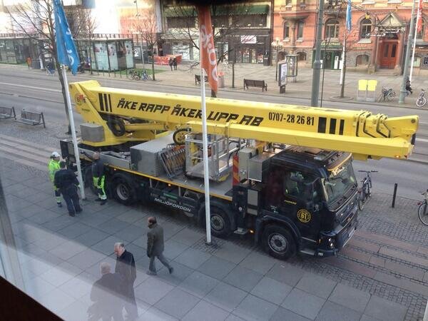 Stor skylift lastbil på en stadsgata, med ett utfällt lyftarm. Flera personer står bredvid.