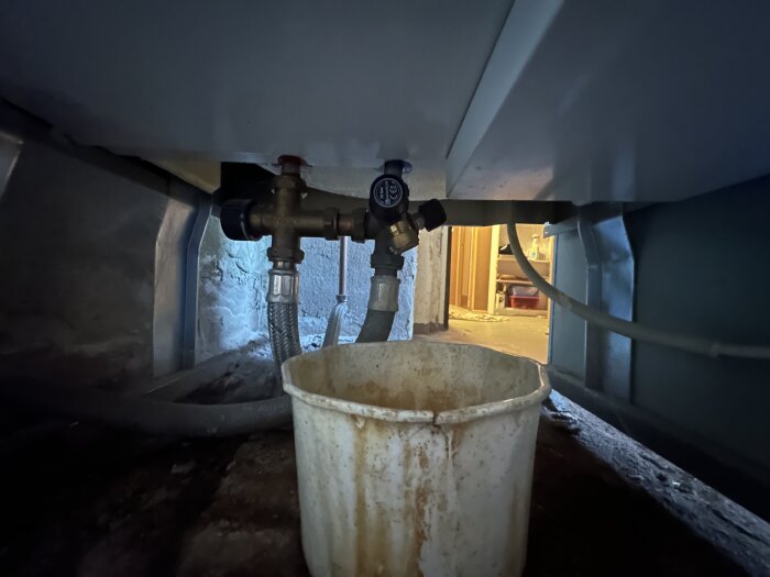 Ventil och kopplingar under varmvattenpanna, med hink placerad nedanför för att fånga upp läckage.