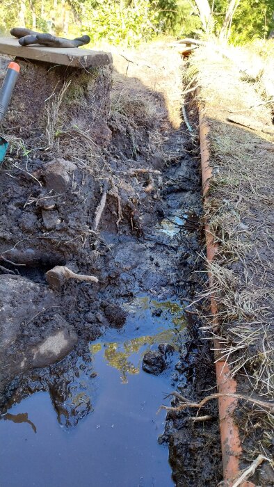 Dike i lerig jord med vatten och ett orange dräneringsrör, del av stugans dräneringsprojekt. Verktyg syns i bakgrunden.