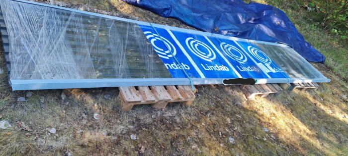 Svarta Lindab-takplåtar inplastade och förvarade på lastpallar under en presenning, liggande på gräs.