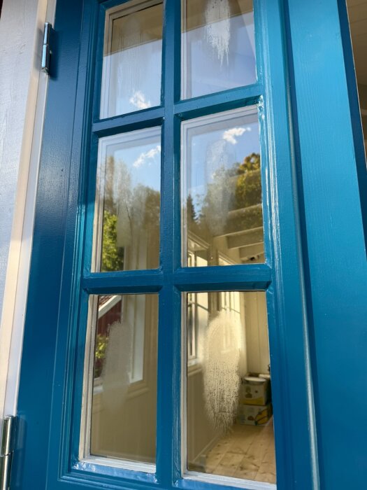 Blåmålad dörr med glasrutor och suddiga märken på glaset; skymtar interiör och träd utanför.